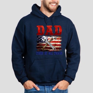 May include: Navy blue hooded sweatshirt with a red and white American flag design. The flag has a wrench and hammer crossed over it. The text "DAD FIXER OF ALL THINGS" is printed below the flag.