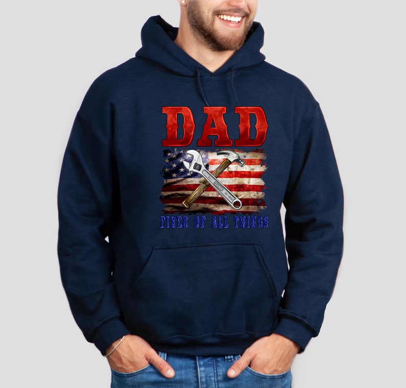 May include: Navy blue hooded sweatshirt with a red and white American flag design. The flag has a wrench and hammer crossed over it. The text "DAD FIXER OF ALL THINGS" is printed below the flag.