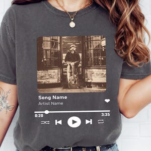 May include: A gray t-shirt with a black and white photo of a person holding a guitar. The photo is in a rectangular frame with a music player interface below it. The interface includes the text "Song Name" and "Artist Name", a play button, and a progress bar.