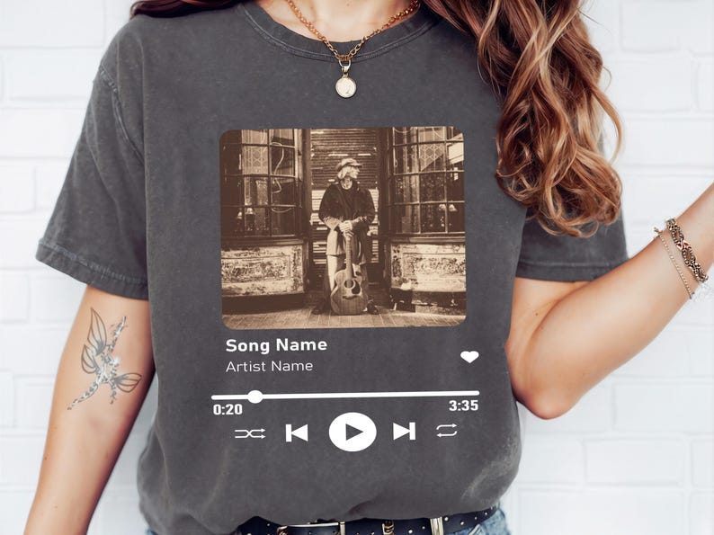 May include: A gray t-shirt with a black and white photo of a person holding a guitar. The photo is in a rectangular frame with a music player interface below it. The interface includes the text "Song Name" and "Artist Name", a play button, and a progress bar.