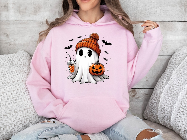 Pu&ograve; includere: Felpa con cappuccio rosa chiaro con un fantasma dei cartoni animati con un berretto arancione, una zucca e una bevanda. Pipistrelli e stelle nere circondano il fantasma, creando un tema di Halloween.