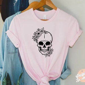 May include: A light pink t-shirt featuring a black skull design adorned with floral accents. The skull has a floral crown and additional flowers at the jawline. The shirt is hanging on a wooden hanger.