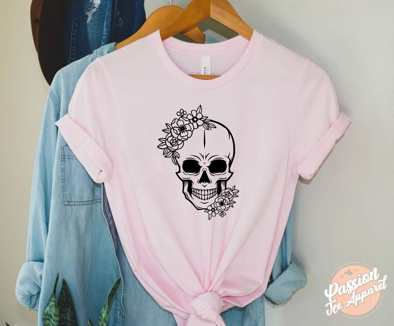 May include: A light pink t-shirt featuring a black skull design adorned with floral accents. The skull has a floral crown and additional flowers at the jawline. The shirt is hanging on a wooden hanger.
