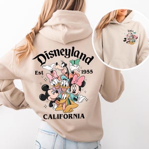 May include: A beige hoodie with a graphic print of Mickey Mouse, Minnie Mouse, Donald Duck, Goofy, and Pluto. The text "Disneyland Est 1955 California" is printed on the back of the hoodie.