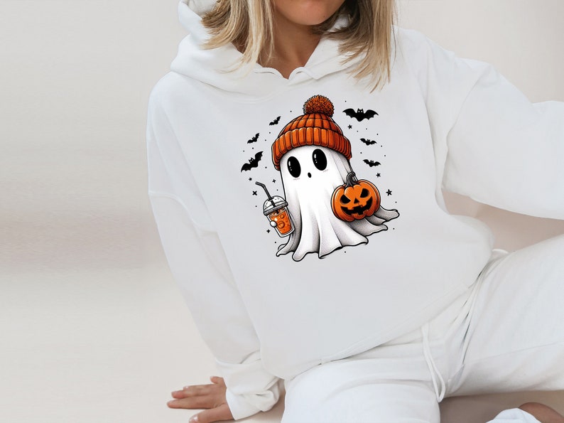 Pu&ograve; includere: Felpa con cappuccio bianca con un fantasma dei cartoni animati che indossa un berretto arancione e tiene in mano una zucca e una bevanda. Pipistrelli e stelle nere circondano il fantasma, creando un tema di Halloween.