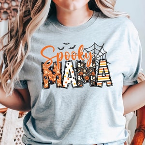以下が含まれることがあります： 「Spooky MAMA」と書かれたライトグレーのTシャツ。 「Spooky」の文字はオレンジ色の筆記体で、「MAMA」はコウモリやカボチャなど、ハロウィーンをテーマにしたパターンでブロック体で書かれています。