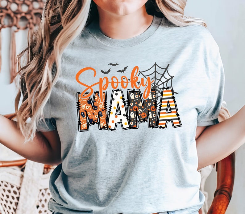 以下が含まれることがあります： 「Spooky MAMA」と書かれたライトグレーのTシャツ。 「Spooky」の文字はオレンジ色の筆記体で、「MAMA」はコウモリやカボチャなど、ハロウィーンをテーマにしたパターンでブロック体で書かれています。