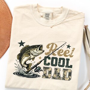 May include: Cream-colored t-shirt with a graphic of a fish jumping out of water, a fishing rod, and the words "Reel COOL DAD" in a mix of fonts and colors. The shirt is made of a soft material and has a crew neck.