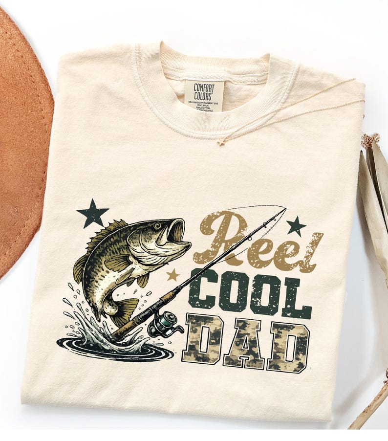 May include: Cream-colored t-shirt with a graphic of a fish jumping out of water, a fishing rod, and the words "Reel COOL DAD" in a mix of fonts and colors. The shirt is made of a soft material and has a crew neck.