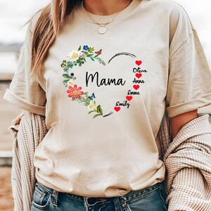 May include: A beige t-shirt featuring a floral heart design with the word "Mama" in the center. The heart is adorned with colorful flowers and butterflies. Names are listed below the heart, with red hearts next to each name.