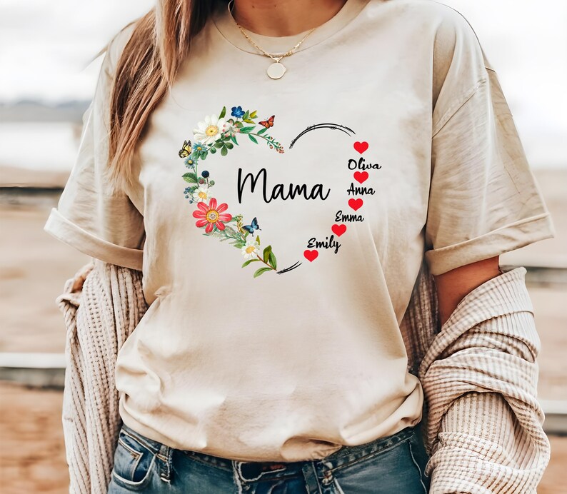 May include: A beige t-shirt featuring a floral heart design with the word "Mama" in the center. The heart is adorned with colorful flowers and butterflies. Names are listed below the heart, with red hearts next to each name.