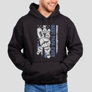 May include: Black hooded sweatshirt with a graphic of fists with names on the knuckles, including "Papa", "Jack", "Jax", and "Sadie". The graphic is set against a blue and white American flag design. The text "Best Papa Ever" is printed vertically on the right side of the graphic.