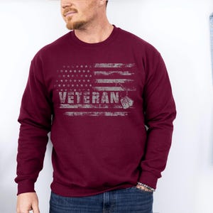 Peut inclure: Un sweat-shirt bordeaux avec un motif de drapeau am&eacute;ricain vieilli et le mot "Veteran" imprim&eacute; dessus.