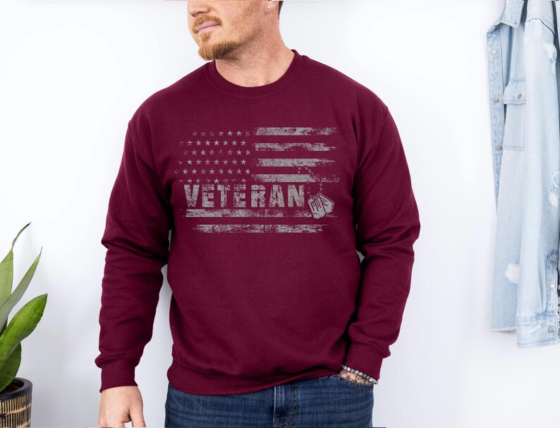 Peut inclure: Un sweat-shirt bordeaux avec un motif de drapeau am&eacute;ricain vieilli et le mot "Veteran" imprim&eacute; dessus.