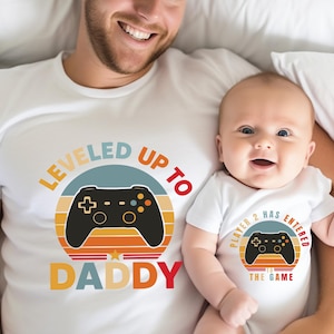 May include: White t-shirts and onesie with matching video game controller graphics. The adult shirt says "LEVELED UP TO DADDY" and the baby's onesie says "PLAYER 2 HAS ENTERED THE GAME". The graphics feature a retro sunset design.