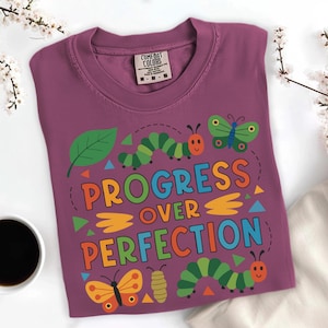 May include: A purple t-shirt with the words "PROGRESS OVER PERFECTION" in colorful letters. The shirt features cartoon butterflies, caterpillars, and a leaf design. The shirt is made of a soft material.