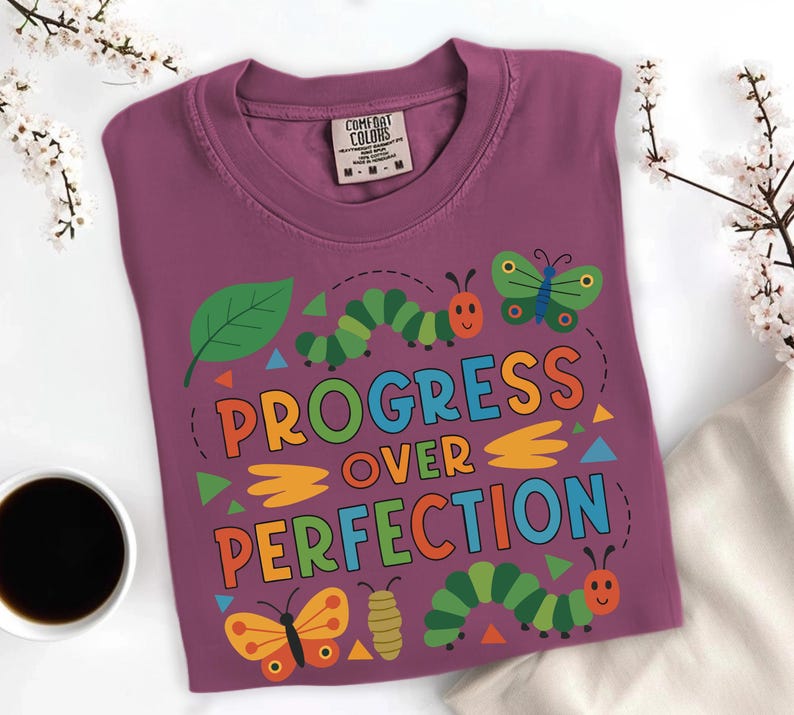 May include: A purple t-shirt with the words "PROGRESS OVER PERFECTION" in colorful letters. The shirt features cartoon butterflies, caterpillars, and a leaf design. The shirt is made of a soft material.