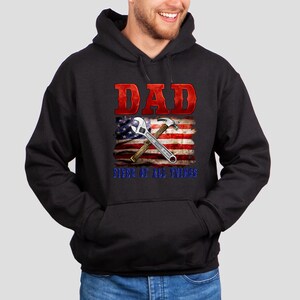 May include: Black hooded sweatshirt with a red, white, and blue American flag design. The flag has a wrench and hammer crossed over it. The text "DAD FIXER OF ALL THINGS" is printed below the flag.