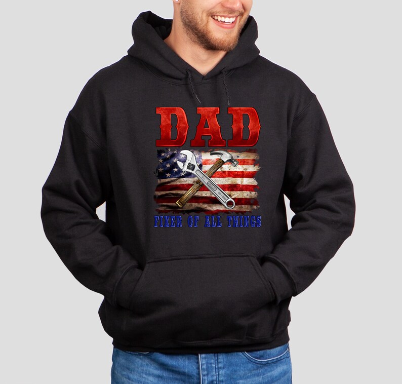 May include: Black hooded sweatshirt with a red, white, and blue American flag design. The flag has a wrench and hammer crossed over it. The text "DAD FIXER OF ALL THINGS" is printed below the flag.