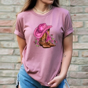 May include: Dusty rose t-shirt featuring a graphic of a pink cowboy hat with a heart, brown cowboy boots, and pink roses. The hat has a fluffy trim. The boots have decorative stitching. The design is centered on the shirt.