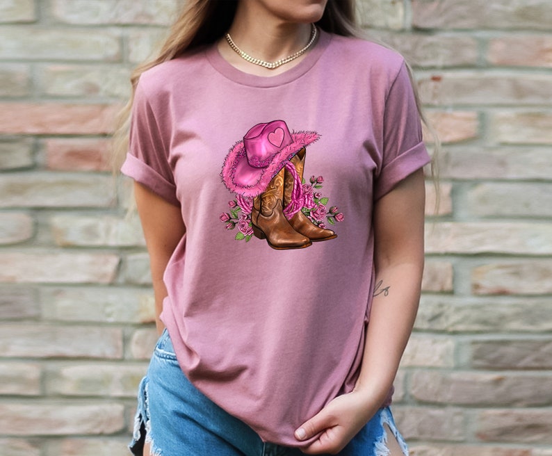 May include: Dusty rose t-shirt featuring a graphic of a pink cowboy hat with a heart, brown cowboy boots, and pink roses. The hat has a fluffy trim. The boots have decorative stitching. The design is centered on the shirt.