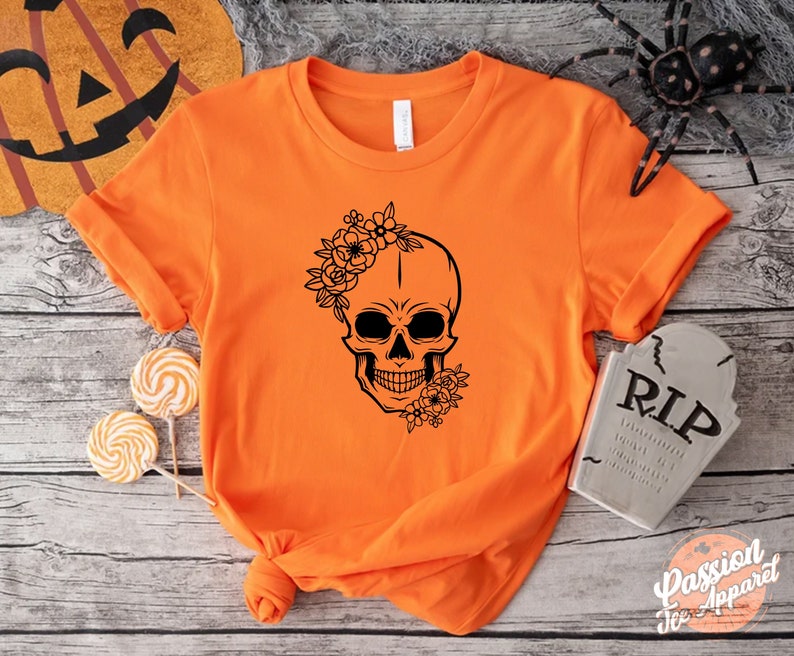 May include: Orange t-shirt featuring a black floral skull design. The skull has flowers on the forehead and jawline. The shirt is a crew neck and has rolled-up sleeves. The shirt is part of a Halloween-themed apparel collection.
