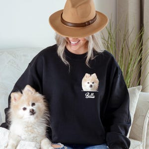 May include: Black sweatshirt featuring a Pomeranian dog portrait with the name "Bella" printed below. The dog is light brown and white. The person in the image is wearing a brown hat.