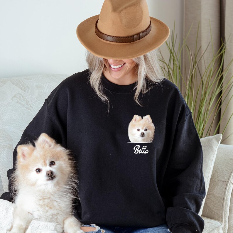 May include: Black sweatshirt featuring a Pomeranian dog portrait with the name "Bella" printed below. The dog is light brown and white. The person in the image is wearing a brown hat.
