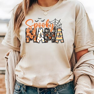 以下が含まれることがあります： オレンジと黒で「Spooky Mama」と書かれたベージュのTシャツ。 「Spooky」の文字はクモの巣のデザインで、「Mama」の文字はカボチャや幽霊などのハロウィーンのパターンで埋められています。 カジュアルで、お祝いの衣服です。