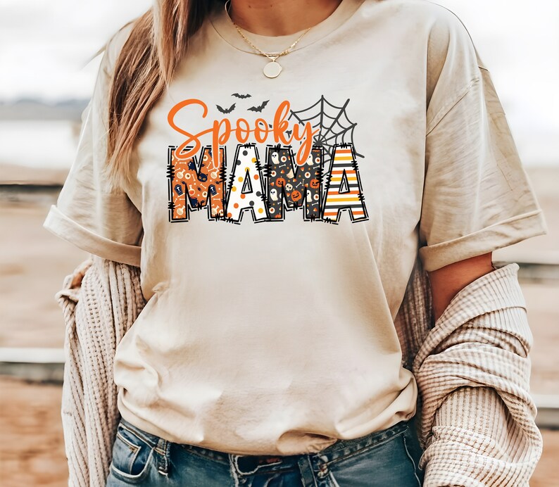 以下が含まれることがあります： オレンジと黒で「Spooky Mama」と書かれたベージュのTシャツ。 「Spooky」の文字はクモの巣のデザインで、「Mama」の文字はカボチャや幽霊などのハロウィーンのパターンで埋められています。 カジュアルで、お祝いの衣服です。