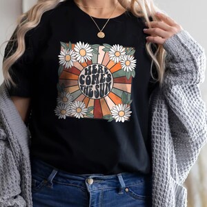 May include: Black t-shirt featuring a retro design with the text "HERE COMES THE SUN" surrounded by daisies and a sunburst pattern in orange, brown, and cream colors. The shirt has a crew neck and short sleeves.