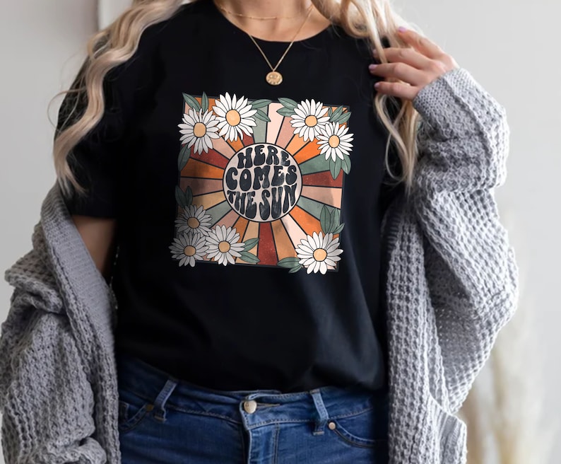May include: Black t-shirt featuring a retro design with the text "HERE COMES THE SUN" surrounded by daisies and a sunburst pattern in orange, brown, and cream colors. The shirt has a crew neck and short sleeves.