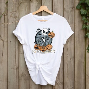 May include: White t-shirt featuring a graphic of Jack Skellington from The Nightmare Before Christmas, holding pumpkins. The design includes bats and the text "THE PUMPKIN KING" on a banner. The shirt is hanging on a wooden hanger.