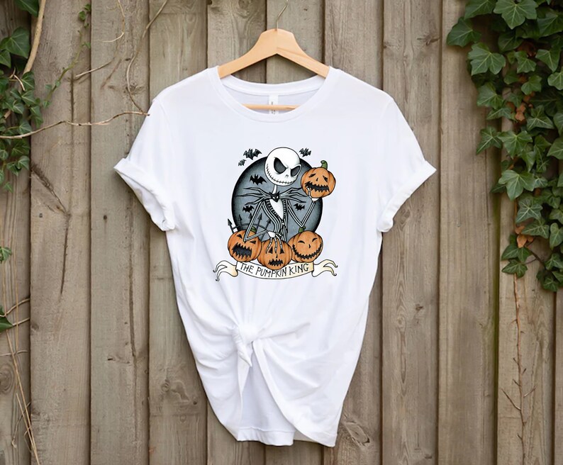 May include: White t-shirt featuring a graphic of Jack Skellington from The Nightmare Before Christmas, holding pumpkins. The design includes bats and the text "THE PUMPKIN KING" on a banner. The shirt is hanging on a wooden hanger.