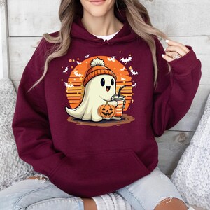 May include: A maroon hoodie featuring a cute cartoon ghost wearing a white beanie and holding a pumpkin and a cup. The ghost is in front of a sunset with bats flying around it.