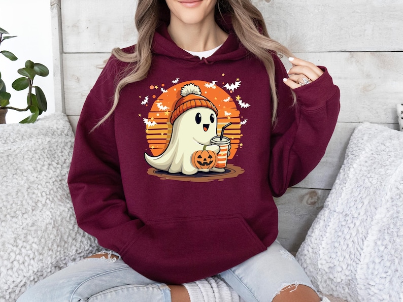 May include: A maroon hoodie featuring a cute cartoon ghost wearing a white beanie and holding a pumpkin and a cup. The ghost is in front of a sunset with bats flying around it.