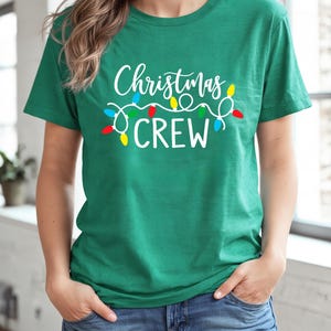 Puede incluir: Una camiseta verde con la frase "Christmas CREW" en blanco y una cadena de luces navide&ntilde;as de colores. La camiseta es de cuello redondo y la lleva una persona. Ideal para Navidad.