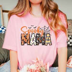 以下が含まれることがあります： オレンジと黒で「Spooky MAMA」と書かれた、明るいピンク色のTシャツ。 「Spooky」という言葉は筆記体で、「MAMA」はハロウィーンをテーマにしたパターンでブロック体で書かれています。 クモの巣とコウモリも特徴です。