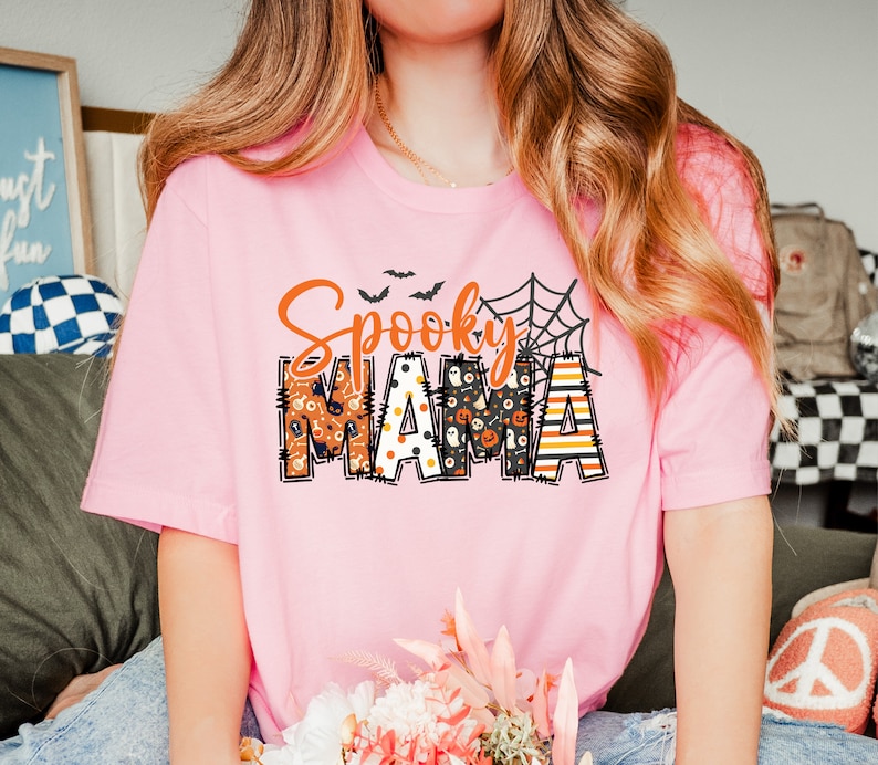 以下が含まれることがあります： オレンジと黒で「Spooky MAMA」と書かれた、明るいピンク色のTシャツ。 「Spooky」という言葉は筆記体で、「MAMA」はハロウィーンをテーマにしたパターンでブロック体で書かれています。 クモの巣とコウモリも特徴です。