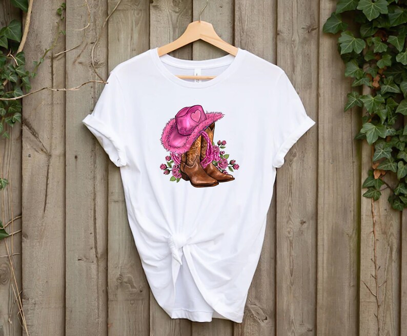 May include: White t-shirt featuring a graphic of a pink cowboy hat with a heart, brown cowboy boots, and pink roses. The design is centered on the shirt, which is hanging on a wooden hanger against a wooden background.