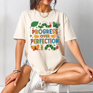May include: Cream-colored t-shirt with the text "PROGRESS OVER PERFECTION" in colorful letters and a whimsical design of butterflies, caterpillars, and a leaf. The shirt is short-sleeved.