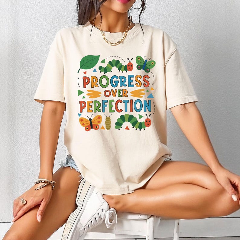 May include: Cream-colored t-shirt with the text "PROGRESS OVER PERFECTION" in colorful letters and a whimsical design of butterflies, caterpillars, and a leaf. The shirt is short-sleeved.