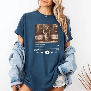 May include: A navy blue t-shirt with a graphic of a music player screen. The screen shows a song title, artist name, and a play button. The song title is "Song Name" and the artist name is "Artist Name".