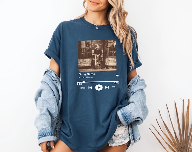 May include: A navy blue t-shirt with a graphic of a music player screen. The screen shows a song title, artist name, and a play button. The song title is "Song Name" and the artist name is "Artist Name".