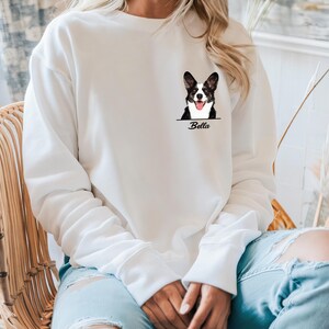 May include: White sweatshirt featuring a black and white dog graphic with the name "Bella" below. The dog graphic is a detailed illustration of a dog's face. The sweatshirt has long sleeves and a crew neck.