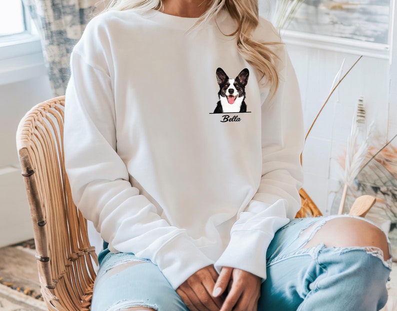 May include: White sweatshirt featuring a black and white dog graphic with the name "Bella" below. The dog graphic is a detailed illustration of a dog's face. The sweatshirt has long sleeves and a crew neck.
