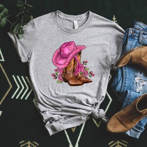May include: A heather gray t-shirt featuring a graphic of a pink cowboy hat with a heart, brown cowboy boots, and pink roses. A pair of blue jeans and brown boots are next to the shirt.