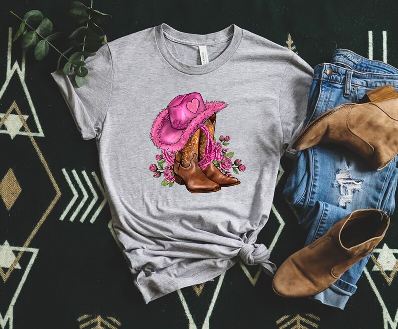 May include: A heather gray t-shirt featuring a graphic of a pink cowboy hat with a heart, brown cowboy boots, and pink roses. A pair of blue jeans and brown boots are next to the shirt.