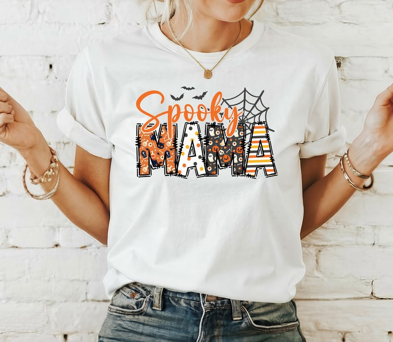 以下が含まれることがあります： オレンジと黒で「Spooky Mama」と書かれた白いTシャツ。「Spooky」は筆記体で、「Mama」はカボチャやコウモリなど、ハロウィーンをテーマにしたパターンでブロック体で書かれています。「Mama」の上にはクモの巣のグラフィックがあります。