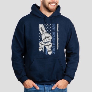May include: Navy blue hooded sweatshirt with a graphic of three fists bumping each other. The fists have the names "Grandpa", "Lily", and "Jack" written on them. The graphic is set against a faded American flag background with the text "Best Grandpa Ever" written vertically.